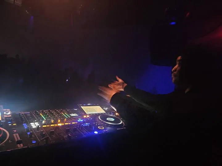 Quentin Hiatus in profile, hand raised over a Pioneer DJ controller in a smoky room